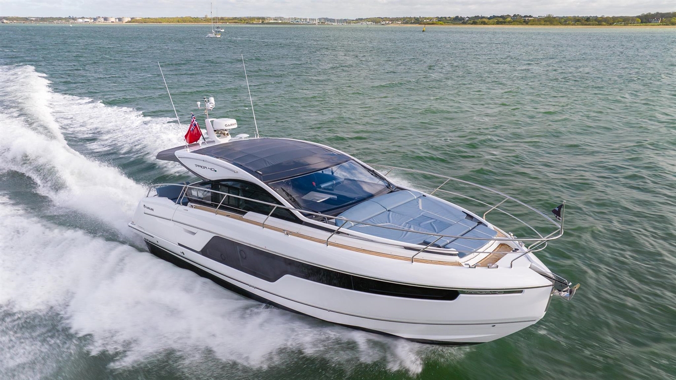 Luxury motor yacht cruising on water, leaving a wake. Features a flag, sleek design, and scenic coastal backdrop.